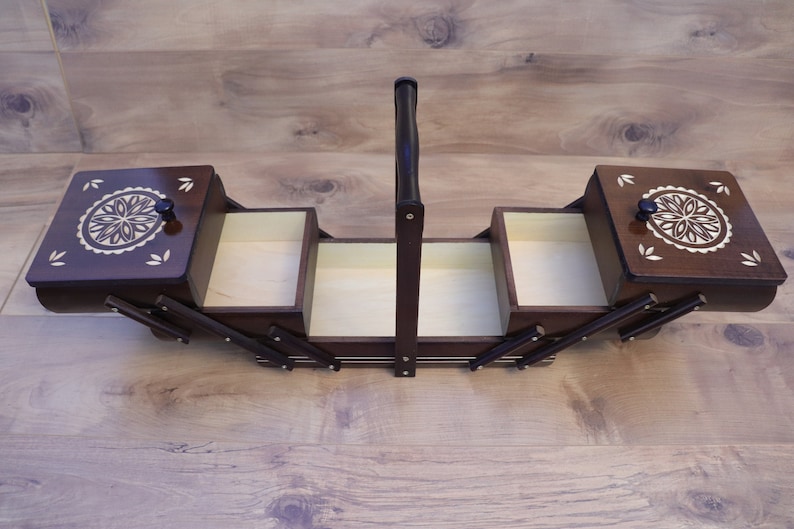 May include: A dark brown wooden sewing box with a cantilevered design, featuring two hinged lids with light-colored floral inlay patterns, a central vertical handle, and multiple open storage compartments with light-colored interior surfaces.
