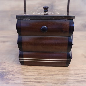 May include: A dark brown wooden sewing box with three stacked drawers, each featuring a rounded front and a black knob. The top lid has a decorative inlay pattern and a small handle. The bottom drawer is accented with two horizontal white lines.