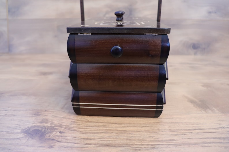 May include: A dark brown wooden sewing box with three stacked drawers, each featuring a rounded front and a black knob. The top lid has a decorative inlay pattern and a small handle. The bottom drawer is accented with two horizontal white lines.