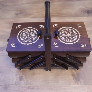 May include: A dark brown wooden sewing box with a tiered accordion-style design and a central carrying handle. The top lid features two circular floral inlays and small leaf-shaped accents. The box has a polished finish and metal hardware.