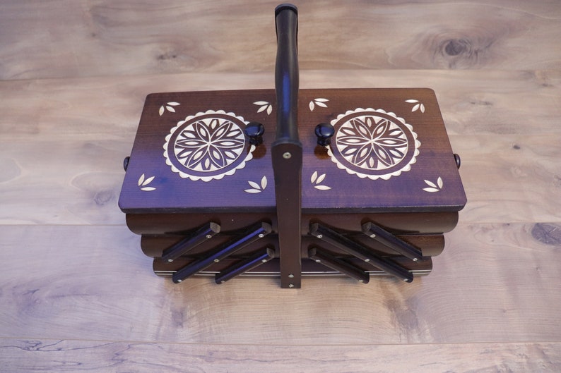May include: A dark brown wooden sewing box with a tiered accordion-style design and a central carrying handle. The top lid features two circular floral inlays and small leaf-shaped accents. The box has a polished finish and metal hardware.