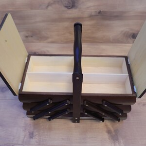 May include: A dark brown wooden sewing box with a cantilevered design and a central carrying handle. The box features two hinged lids that open to reveal a light-colored interior compartment with a central divider for organized storage.