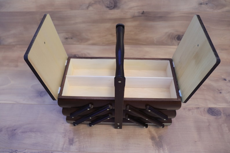 May include: A dark brown wooden sewing box with a cantilevered design and a central carrying handle. The box features two hinged lids that open to reveal a light-colored interior compartment with a central divider for organized storage.