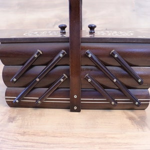 May include: A dark brown wooden sewing box with a tiered accordion-style design. The box features a central vertical handle, two small knobs on the top lids, and metal hinges connecting the three horizontal storage compartments together.
