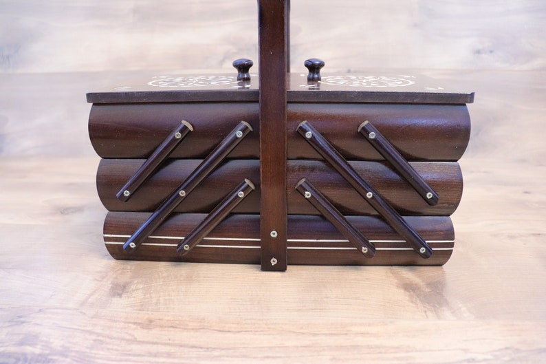 May include: A dark brown wooden sewing box with a tiered accordion-style design. The box features a central vertical handle, two small knobs on the top lids, and metal hinges connecting the three horizontal storage compartments together.