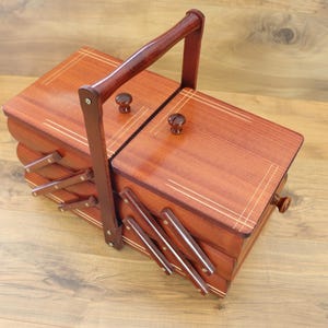 May include: A reddish-brown wooden sewing box with a tiered accordion-style design, featuring two top lids with small round knobs, a tall central carrying handle, and light-colored inlaid line details on the lids and side panels.