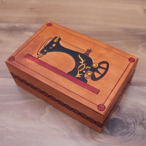 May include: A rectangular wooden box with a light brown finish featuring a dark blue and yellow illustration of a vintage sewing machine on the lid. The box is decorated with a red border, small red circles at the corners, and a red scalloped trim.