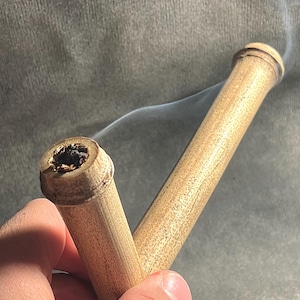 May include: A handcrafted bamboo pipe with a V-shaped design. The pipe is light brown with visible wood grain and a dark bowl. Wisps of smoke are rising from the pipe, suggesting recent use. Held in a hand.