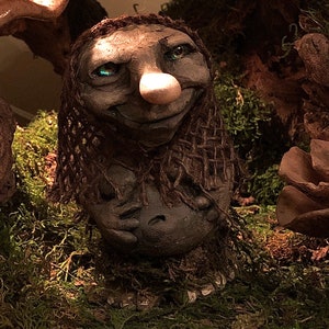 May include: A small, handmade troll figurine with a large, bulbous nose and green eyes. The troll is made of a dark brown material and has a mossy green base.