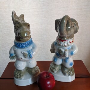 May include: Two ceramic figurines, one of a rhino and one of an elephant, both wearing blue and white pajamas with red accents. The figurines are standing on a wooden surface with a red apple in front of them.