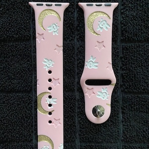 May include: A light pink silicone watch strap featuring a repeating pattern of gold glitter crescent moons, white embossed stars, and small white embossed rabbit faces. The strap includes a silver metal pin closure and standard attachment connectors.