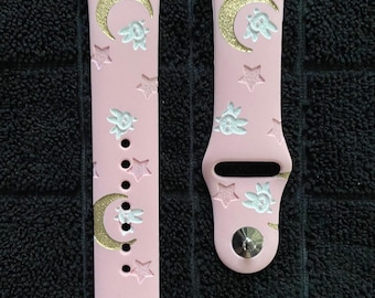Può includere: Un cinturino per orologio in silicone rosa chiaro con un motivo ripetuto di lune crescenti glitterate dorate, stelle bianche in rilievo e piccoli volti di coniglio bianchi in rilievo, dotato di chiusura a perno in metallo argentato.