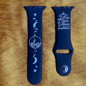 May include: A black silicone watch strap featuring white printed designs. One piece displays a mountain range inside a circle with crescent moons and stars. The other piece features the text: To the stars who listen & the dreams that are answered.