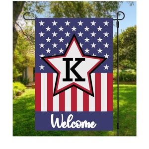 May include: A patriotic garden flag featuring a blue background with white stars and red and white stripes. A large white star with a red outline and the letter "K" is centered. The word "Welcome" is written in white script.