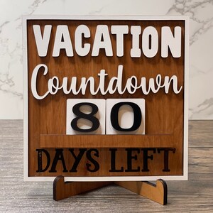 A wooden countdown sign with white lettering that reads 'Vacation Countdown 80 Days Left'. The sign is standing on a small wooden stand.