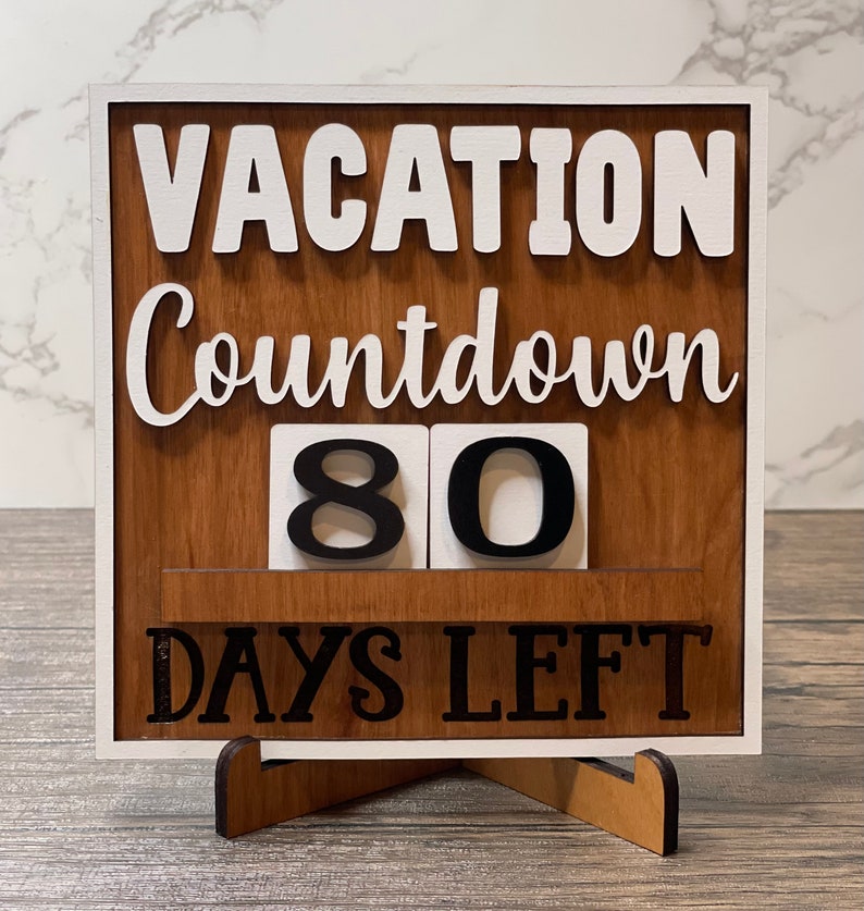 A wooden countdown sign with white lettering that reads 'Vacation Countdown 80 Days Left'. The sign is standing on a small wooden stand.