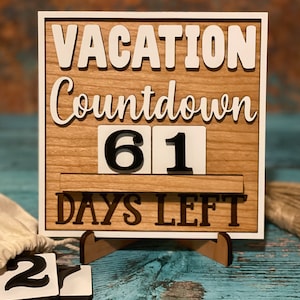 A wooden countdown sign with white lettering that reads 'Vacation Countdown 61 Days Left'. The sign is on a stand and has a white background with a wood grain texture. The numbers 61 are displayed on the sign.