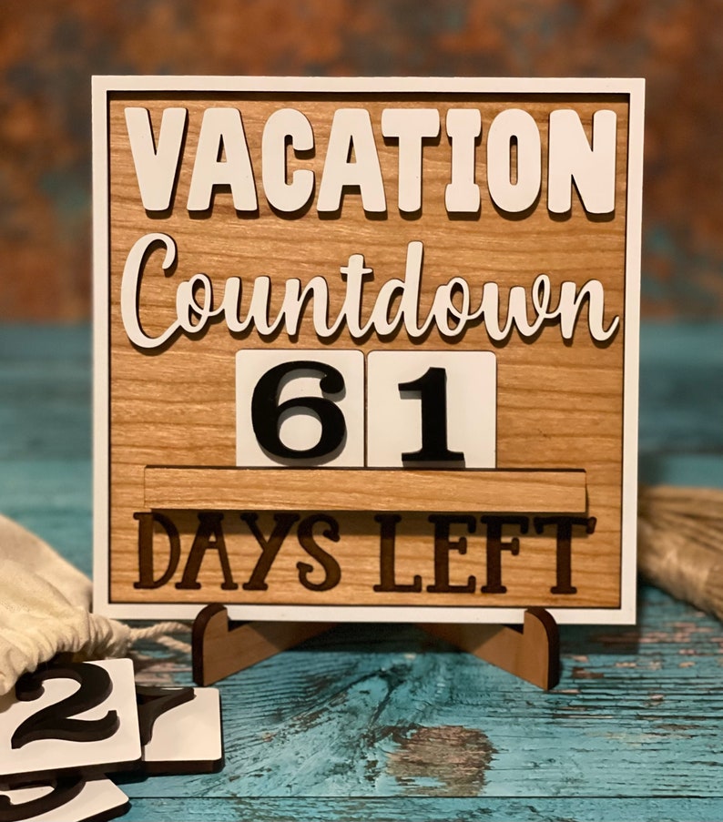 A wooden countdown sign with white lettering that reads 'Vacation Countdown 61 Days Left'. The sign is on a stand and has a white background with a wood grain texture. The numbers 61 are displayed on the sign.
