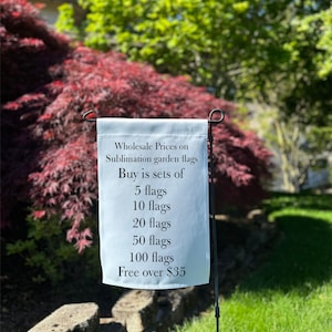 May include: A white garden flag displays wholesale prices for sublimation garden flags. The text lists sets of 5, 10, 20, 50, and 100 flags. The flag is mounted on a black metal stand, set against a backdrop of green grass and red and green foliage.