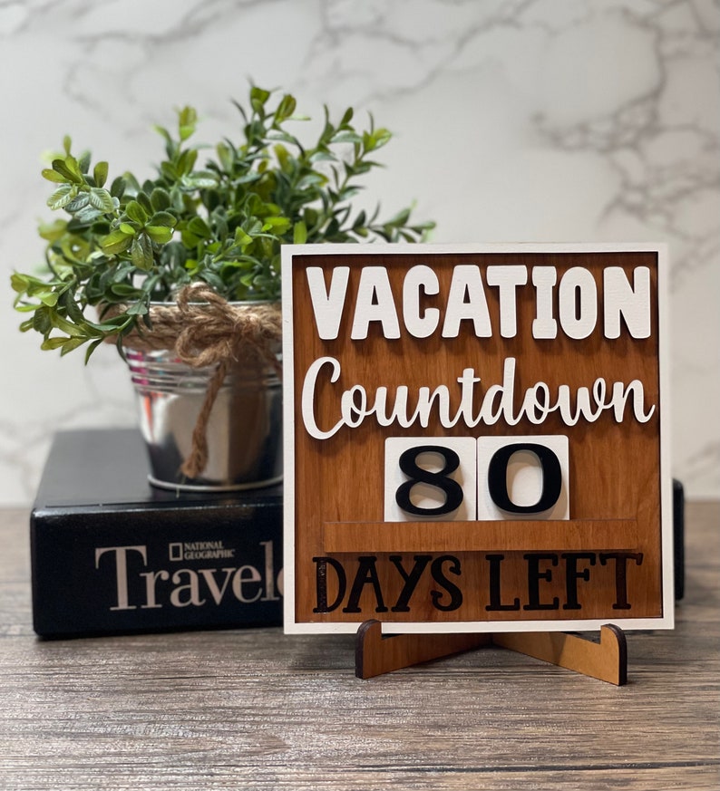 A wooden countdown sign with the text 'Vacation Countdown 80 Days Left' in white and black lettering. The sign is standing on a small wooden stand