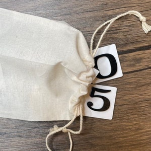 A cream-colored drawstring bag rests on a wood surface with two white number tiles partially spilling out, displaying the numbers 0 and 5 in black raised lettering.