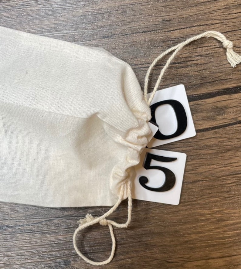 A cream-colored drawstring bag rests on a wood surface with two white number tiles partially spilling out, displaying the numbers 0 and 5 in black raised lettering.