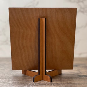 Wooden display stand shown from the back, featuring a natural wood grain panel supported by an interlocking wooden base on a rustic tabletop.