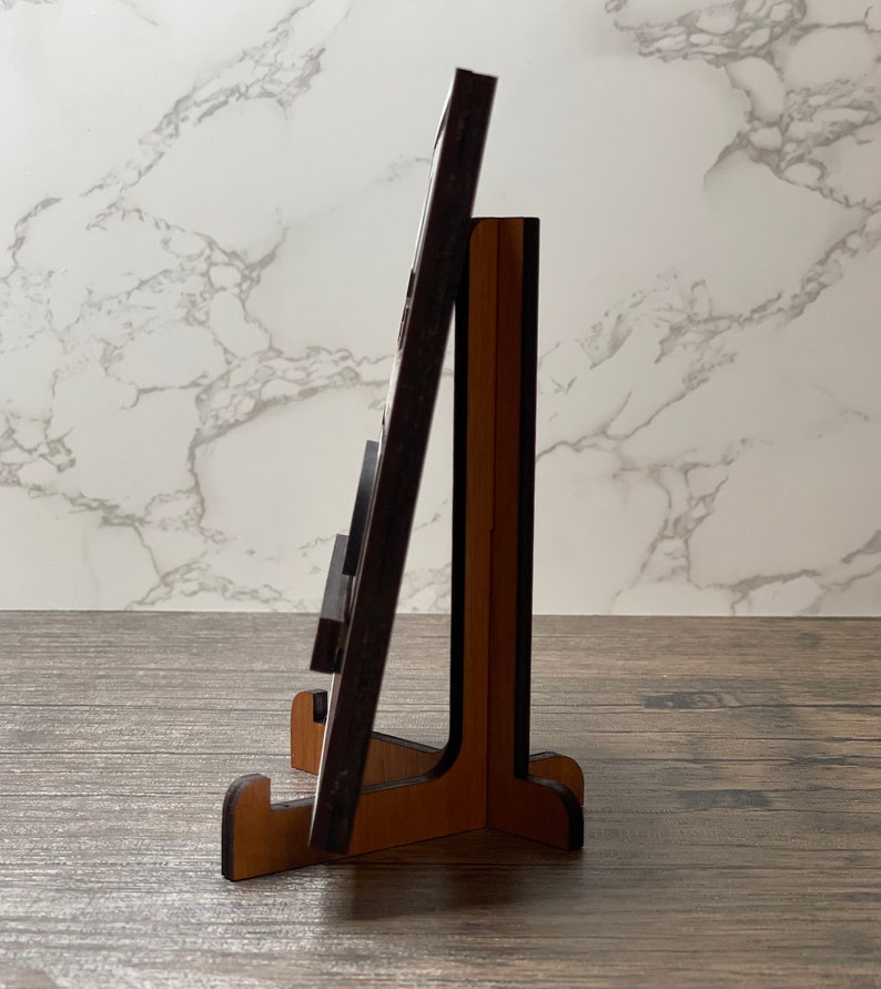 Side view of a wooden display stand holding a thin wooden sign, shown on a rustic tabletop with a marble-patterned background