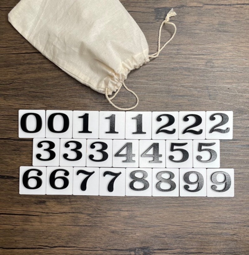 Set of white number tiles arranged from 0 to 9 with multiples of each digit, displayed on a wood surface alongside a drawstring storage bag.