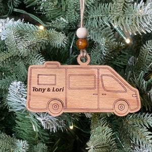 May include: Wooden Christmas ornament shaped like a camper van with the names "Tony & Lori" engraved on the side. The ornament is hanging from a tree with brown and white beads.