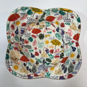 May include: A white fabric bowl with a beach-themed print. The print features various beach-related icons, including seashells, palm trees, sunglasses, and the text "Sea & Sand", "Beach Vibes", and "Hello Sunshine".