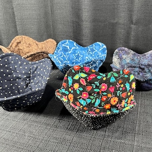 May include: Five fabric bowl cozies in various patterns. One is navy with white polka dots, another is black with a floral design, and a third is blue with white stars. The other two are brown and blue with a galaxy design.