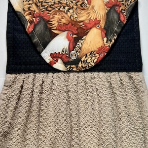 May include: A brown kitchen towel with a black pocket featuring a colorful rooster print. The pocket has a black border and a white button.