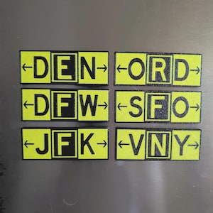 May include: Six rectangular magnets with a bright lime green background and black text. Each magnet features a three-letter airport code: DEN, ORD, DFW, SFO, JFK, and VNY. Each code is flanked by black arrows pointing left and right.