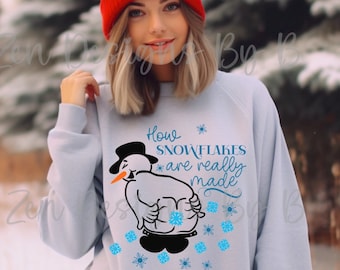 Op de afbeelding: Een lichtblauw sweatshirt met een afbeelding van een sneeuwman met een oranje wortelneus en een zwarte hoge hoed. De sneeuwman bukt voorover met blauwe sneeuwvlokken die erachter vandaan komen. De tekst luidt How SNOWFLAKES are really made.