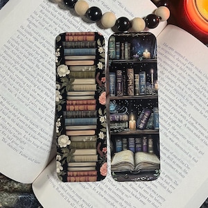 May include: Two bookmarks featuring a stack of books with a floral design and a bookshelf with a glowing candle and an open book. The bookmarks are black with a dark, vintage aesthetic.