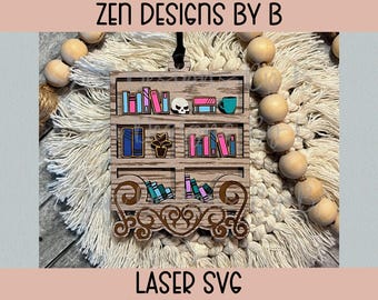 May include: A laser-cut wooden bookshelf ornament featuring three shelves filled with colourful miniature books, a skull, a potted plant, and a mug. The base has an ornate scrollwork design. The text ZEN DESIGNS BY B LASER SVG is visible.
