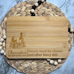 May include: A wooden cutting board with a laser-engraved design of a wine glass, grapes, cheese, and a bottle of wine. The text "Charcuterie" is engraved below the design, along with the definition "French word for cheese and other fancy shit."