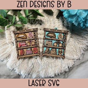 May include: Two wooden shelf ornaments with various items, including books, handcuffs, and a lock. The shelves have the words "GOOD GIRL" and "SMUT FUND" on them. The ornaments are laser cut and sit on a cream-colored surface.