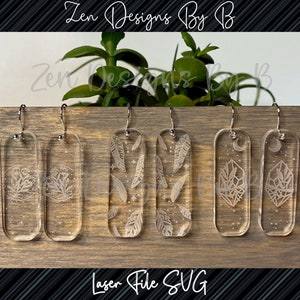 May include: Three pairs of clear rectangular acrylic earrings with silver-toned hooks. The first pair features etched floral designs, the second pair displays a pattern of feathers and stars, and the third pair shows a crystal and moon motif.