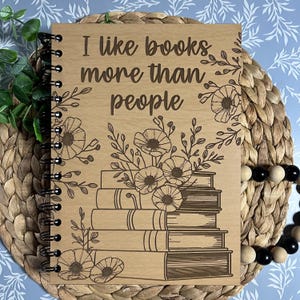 May include: A spiral-bound notebook with a light brown wooden cover featuring the engraved text I like books more than people. The design includes a stack of books and floral illustrations in dark brown, with black metal binding rings.