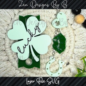 May include: A green four-leaf clover with the word "lucky" written in black cursive. A green shamrock, a silver chain with a horseshoe and a heart charm, and a wooden bead are also included. The items are on a white background.