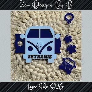 May include: Blue and white wooden cutout of a Volkswagen van with the name "Bethanie" written on the bottom. The van is attached to a blue and white charm with a flower design. The charm is attached to a silver chain with a ring at the top.
