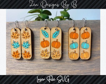 May include: Three pairs of wooden earrings with different fall-themed designs. The first pair features orange and blue leaves, the second pair features orange pumpkins and blue leaves, and the third pair features orange pumpkins and blue pumpkins.