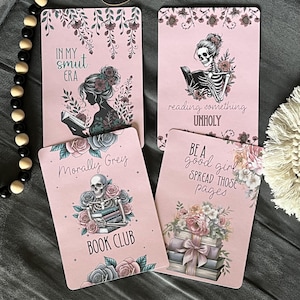 May include: Four rectangular pink bookmarks featuring floral illustrations and skeletons. Text includes: In My Smut Era, reading something UNHOLY, Morally Grey BOOK CLUB, and BE A good girl & SPREAD THOSE pages. Each card displays unique book-themed art.