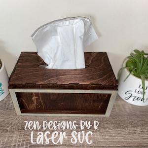 以下が含まれることがあります： 白いティッシュボックスが入った、濃い茶色の木製のティッシュボックスカバー。ティッシュボックスカバーは、縁に白い縁取りが施されています。ティッシュボックスカバーは木製の表面の上に置かれています。ティッシュボックスカバーの下の表面には、「zen Designs BY B Laser - SVG」という文字が印刷されています。