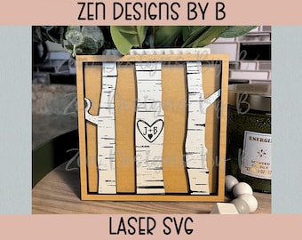 May include: A square wooden sign featuring three white birch tree trunks with black horizontal markings. The center trunk displays a carved heart containing the letters J+B. The text ZEN DESIGNS BY B and LASER SVG appears at the top and bottom.