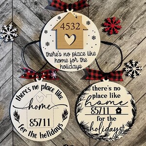 May include: Three wooden holiday ornaments with address numbers and the phrase "there's no place like home for the holidays." Each ornament has a black string for hanging and a red and black plaid bow. Decorative snowflakes are also present.