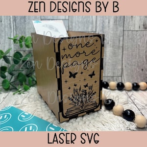 Könnte beinhalten: Eine hellbraune Holzbox mit einem lasergeschnittenen Design auf der Vorderseite. Das Design zeigt den Text "one more page" über einem offenen Buch mit Blumen, Schmetterlingen und Sternen. Die Box hat sichtbare Verbindungsstücke.
