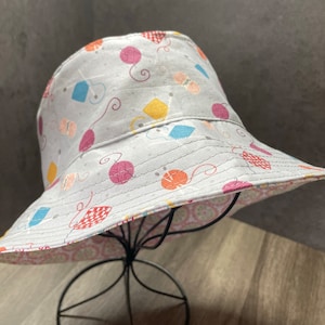 May include: A white bucket hat with a pink, yellow, and blue yarn ball pattern. The hat has a pink and white patterned lining.